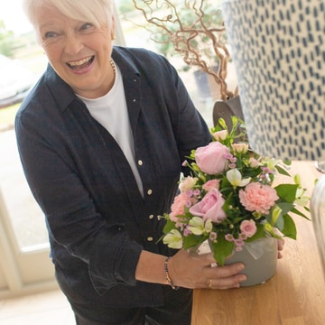 A smiling woman places a beautiful pink and green florist-choice arrangement on a wooden table near a bright window