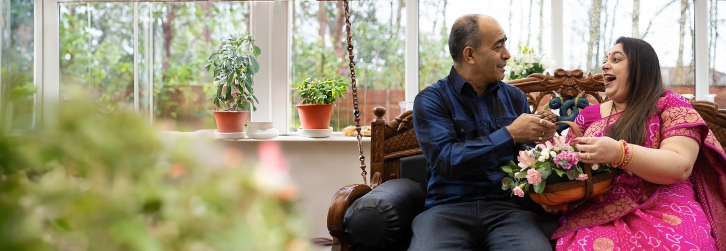 A couple sit together smiling as the man presents a basket of pink and white flowers to the woman.