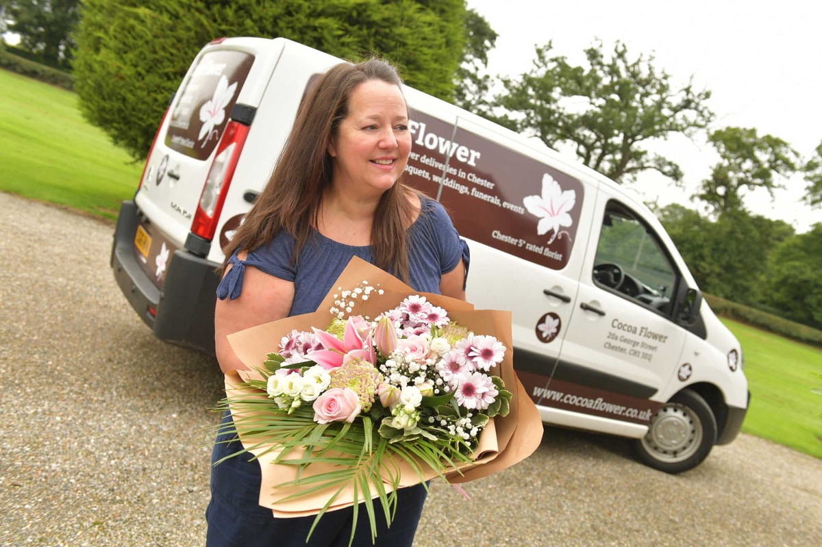 Woman florist with delivery van behind her holding a pink wrap of flowers she's delivering.