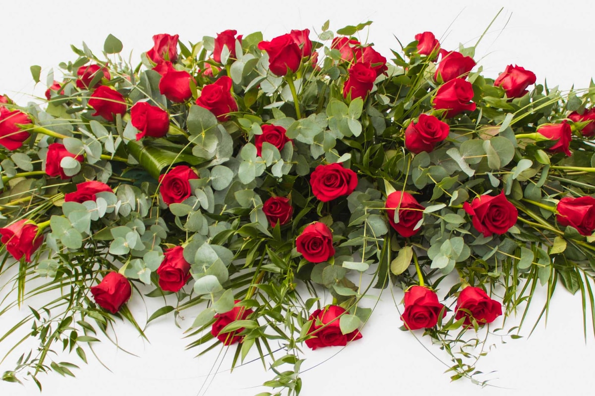 Close-up of a large funeral spray with numerous red roses and green foliage on a white background.