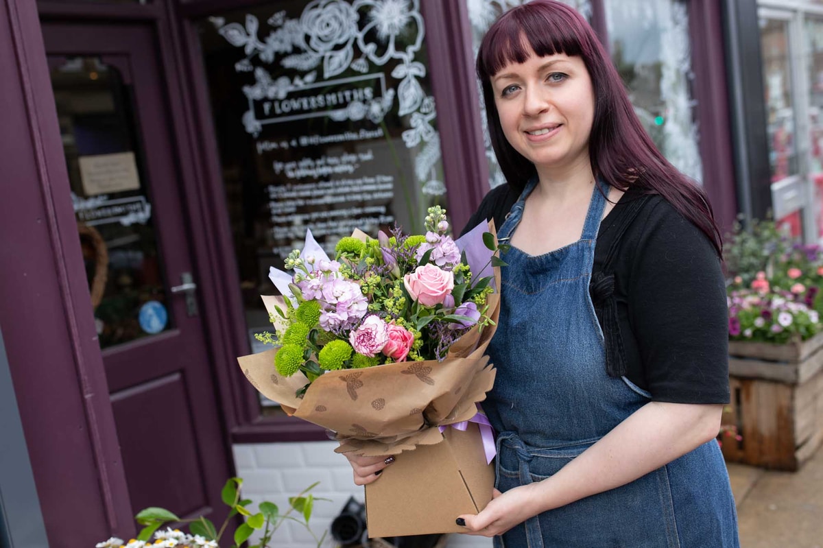 Florist woman in a denim apron in front of shop smiling, holding colourful hand-tied in a box.