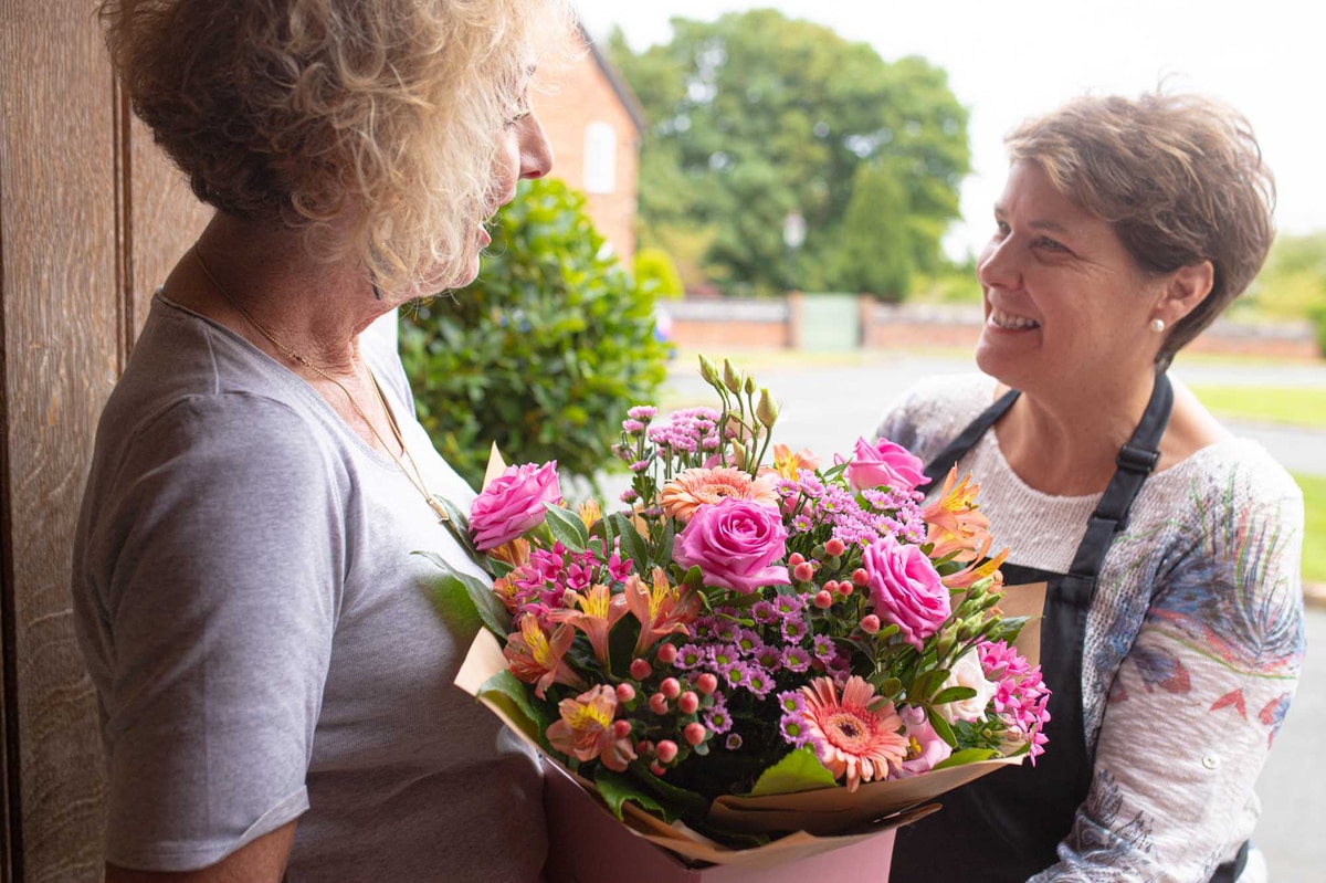 Women in happy conversation with a florist thats handing her a delivery of pink and peach themed flowers.