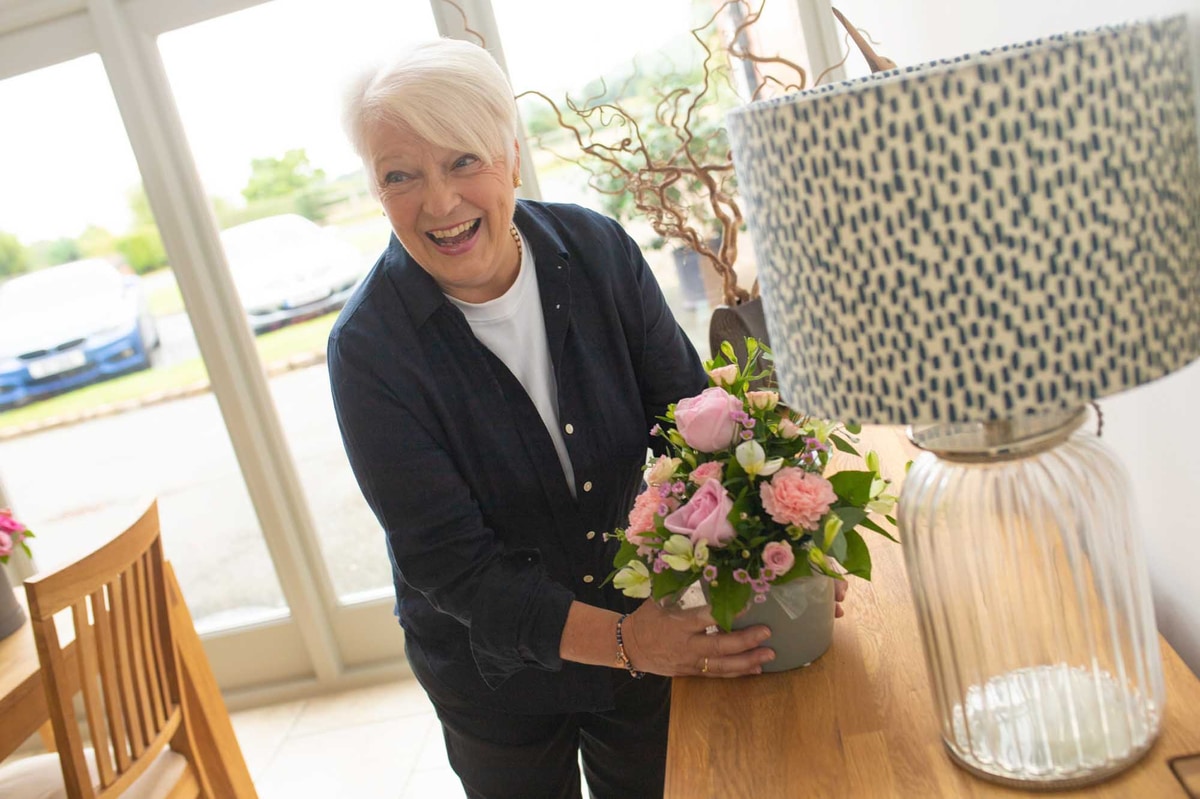 Older woman laughing and looking at someone whilst placing a pot of flowers on a sideboard.